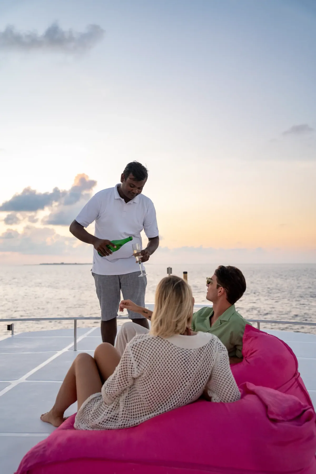 Niva Velassaru Maldives, activity, South Malé Atoll, Maldives, Sunset cruise on the property yacht: couple lounging on a hot-pink bean-bag deck cushion, crew member pouring champagne for them, the sun dropping to the open-ocean horizon, the post-sunset gradient brushing the clouds.