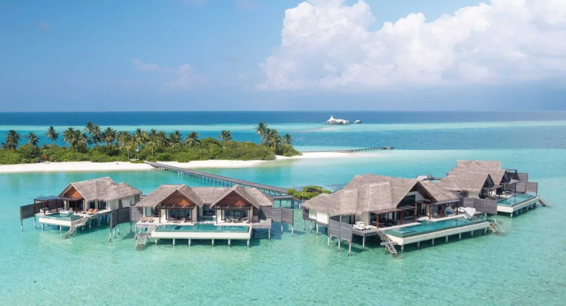 Niyama Private Islands Maldives, aerial, Dhaalu Atoll, Maldives, Mid-altitude aerial of the over-water Crescent Pool Suites cluster, large two-bedroom configurations with thatched-roof pavilions and private infinity pools on timber decks, the signature white tent-roof Edge pavilion visible offshore at upper-right, the jetty bridging back to the palm-canopy main island.