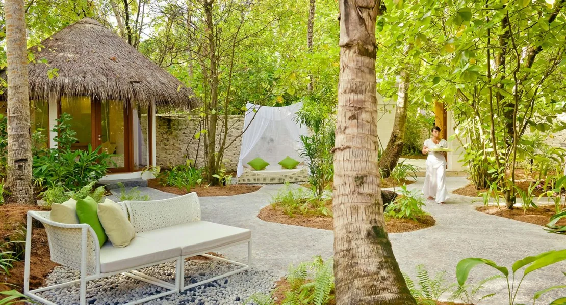 Niyama Private Islands Maldives, spa, Dhaalu Atoll, Maldives, Drift Spa garden setting, thatched-roof spa pavilion at left, white-canvas garden bed with sheer netting, white woven sofa at the path edge, lush jungle and palm canopy throughout, a therapist approaching with a tray at right.