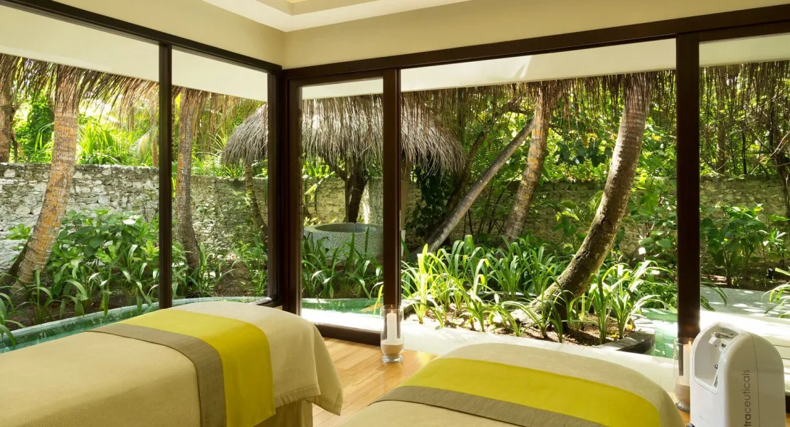 Niyama Private Islands Maldives, spa, Dhaalu Atoll, Maldives, Drift Spa couple treatment room, twin massage tables with mustard-yellow throws, floor-to-ceiling glass on two sides looking out to the jungle garden, an outdoor thatched relaxation pavilion and small plunge pool visible through the windows.