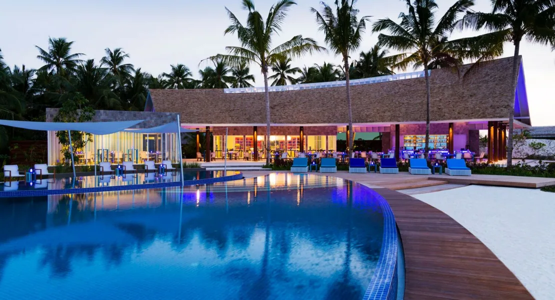 Niyama Private Islands Maldives, dining, Dhaalu Atoll, Maldives, Main pool and the curved-roof main pavilion at twilight, a large curved infinity pool reflecting the blue interior illumination of the pavilion, palm canopy framing the lit pavilion windows, sun loungers along the timber deck.