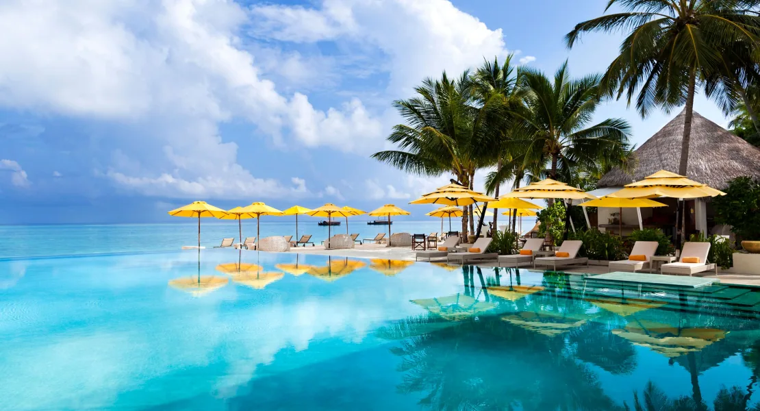 Niyama Private Islands Maldives, setting, Dhaalu Atoll, Maldives, Main infinity pool with yellow umbrellas and sun loungers along the timber deck, palm canopy framing at right, a thatched-roof poolside pavilion at the far edge, lagoon visible beyond the infinity edge.
