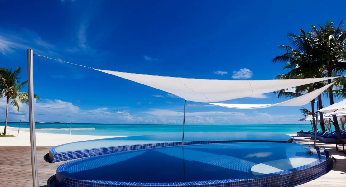 Niyama Private Islands Maldives, setting, Dhaalu Atoll, Maldives, Circular beach club infinity pool with a triangular white sail-cloth shade tensioned overhead, blue-mosaic-tile pool bowl, sand and timber deck around the perimeter, lagoon stretching to the horizon beyond the infinity edge.
