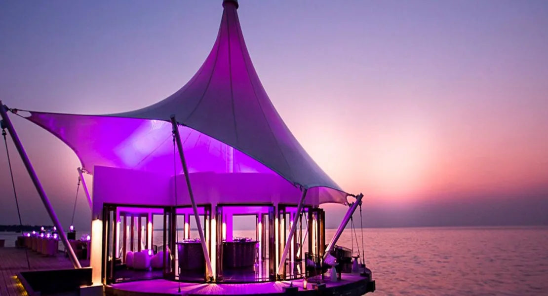 Niyama Private Islands Maldives, dining, Dhaalu Atoll, Maldives, Edge over-water restaurant at twilight, the property's signature white tensile-fabric tent roof on guy-cables illuminated from within in purple, the round overwater pavilion floor with surrounding deck floats above the lagoon, pink-orange horizon at sunset.
