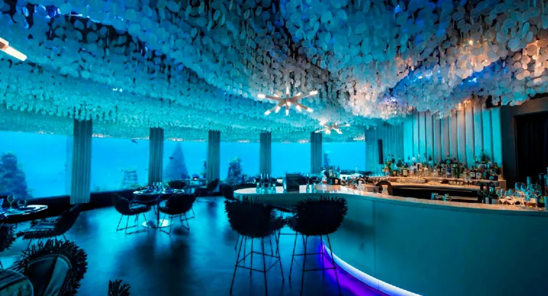 Niyama Private Islands Maldives, dining, Dhaalu Atoll, Maldives, Subsix underwater nightclub interior, the country's only standalone underwater dining venue at approximately six metres below sea level, signature capiz-shell ceiling and starburst chandelier, curved clam-inspired bar with black anemone-style chairs, panoramic underwater windows with the surrounding reef visible through the blue glow.