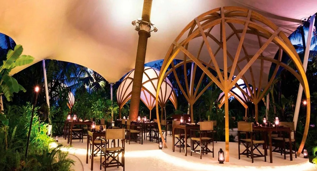Niyama Private Islands Maldives, dining, Dhaalu Atoll, Maldives, Tribal restaurant, distinctive wooden ribbed dome pavilions paired with white tensile-fabric tent canopies (the property's signature dining architecture), sand floor dining tables, lanterns lit at night, palms framing the perimeter.