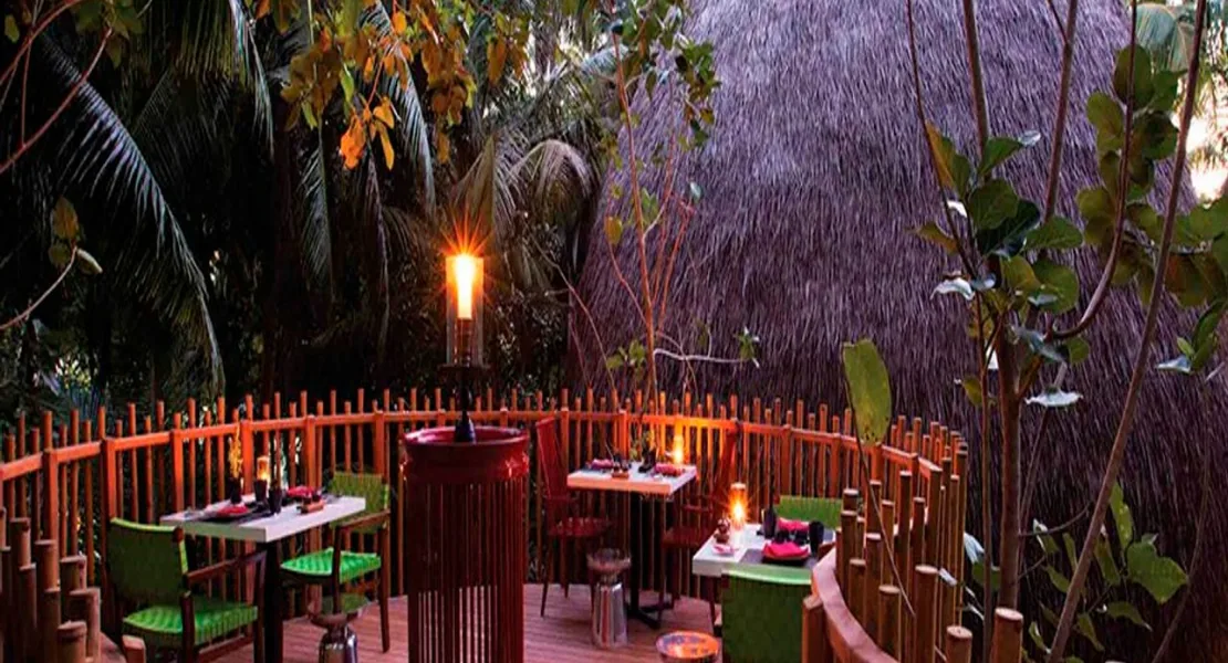 Niyama Private Islands Maldives, dining, Dhaalu Atoll, Maldives, Nest treehouse-style restaurant, raised bamboo-balustrade dining deck with green-cushioned wicker chairs around small tables, lit bamboo torch and tabletop candles, woven palm-leaf thatched feature wall, tropical foliage framing the open-air pavilion.