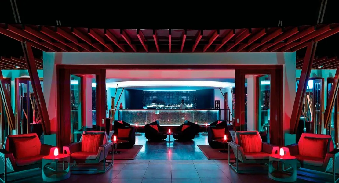 Niyama Private Islands Maldives, dining, Dhaalu Atoll, Maldives, Fahrenheit bar lounge, geometric red-cedar ceiling beams over a moody red-and-black interior, modern lounge seating with red cushions and small red side tables, the bar with backlit bottles at the center distant.