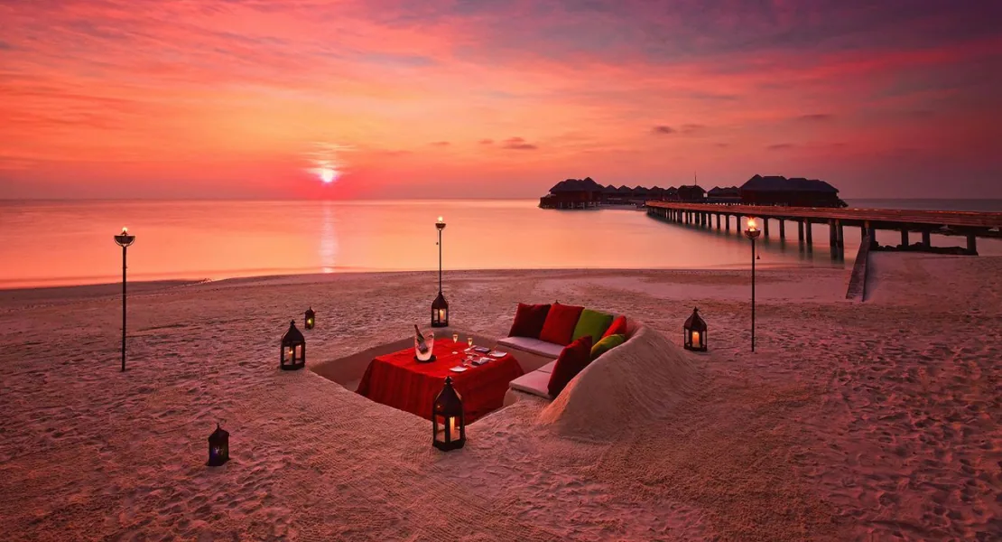 Niyama Private Islands Maldives, dining, Dhaalu Atoll, Maldives, Beach destination dining at sunset, a sunken dining table-in-sand setup with red linen, cushions, and lanterns, overwater villa row and jetty extending to the horizon at right, pink-red sunset stretched across the lagoon.