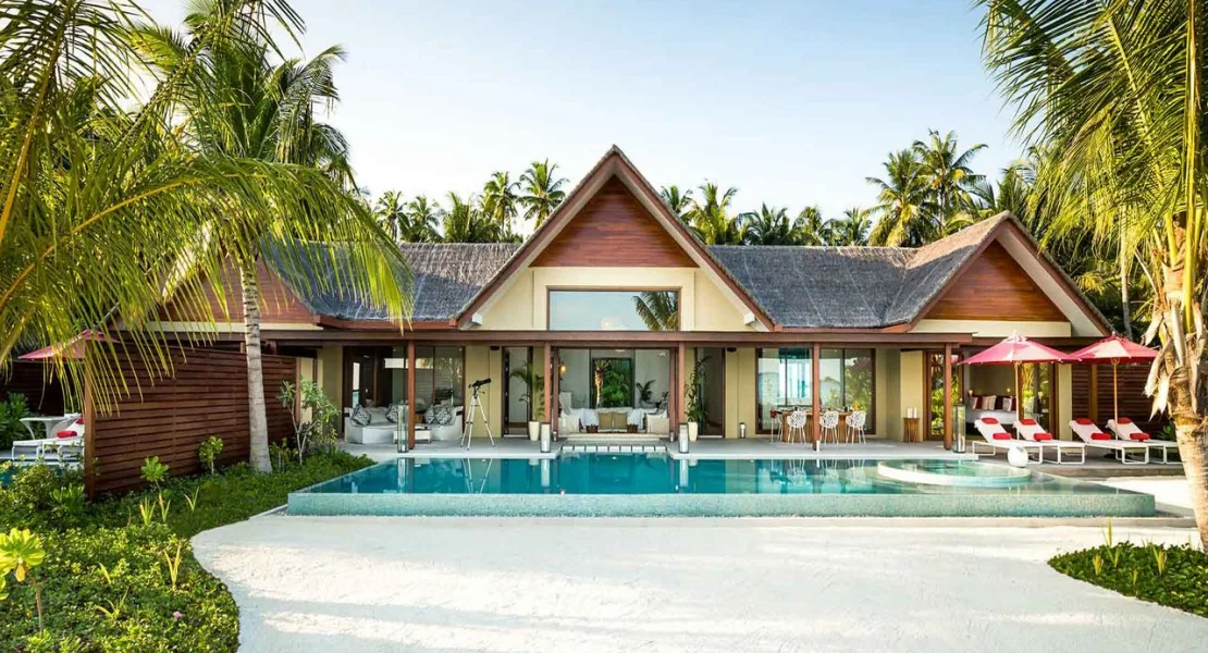 Niyama Private Islands Maldives, villa beach, Dhaalu Atoll, Maldives, Two-Bedroom Beach Pool Villa exterior, a symmetric twin-peak gabled-roof pavilion with thatched eaves, a large rectangular infinity pool with sun loungers and red umbrellas, palm canopy framing both sides, indoor lounge visible through floor-to-ceiling glass.