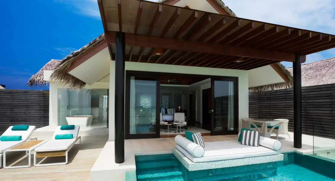 Niyama Private Islands Maldives, villa overwater, Dhaalu Atoll, Maldives, Water Pool Villa exterior deck, a wide pavilion with twin thatched-roof wings, timber-and-mesh pergola over the deck, central infinity pool with a sunken daybed and pop-color cushions, twin sun loungers with aqua throws, indoor lounge and freestanding bathtub visible through the glass sliders.