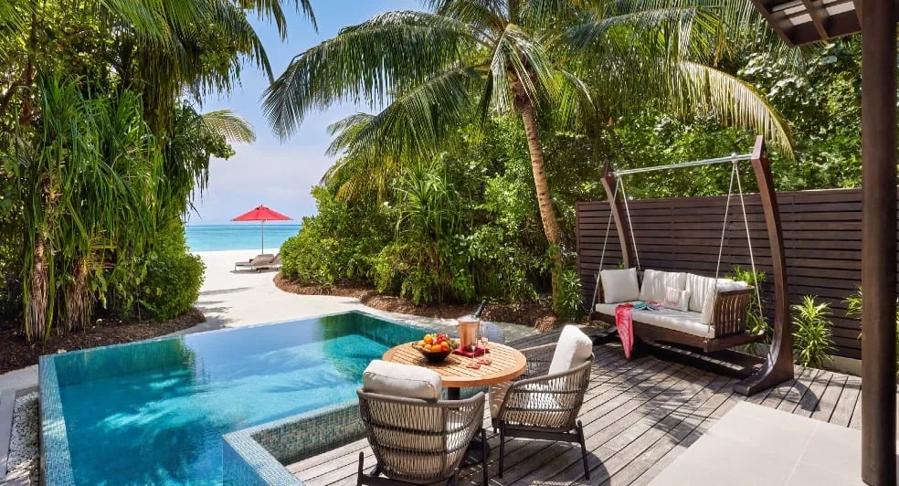 Niyama Private Islands Maldives, villa beach, Dhaalu Atoll, Maldives, Beach Pool Villa terrace, a rectangular plunge pool, swing chair, round wooden table with fresh fruit and chilled drink, palm canopy framing the beach view with a red parasol pinned in the sand in the background.