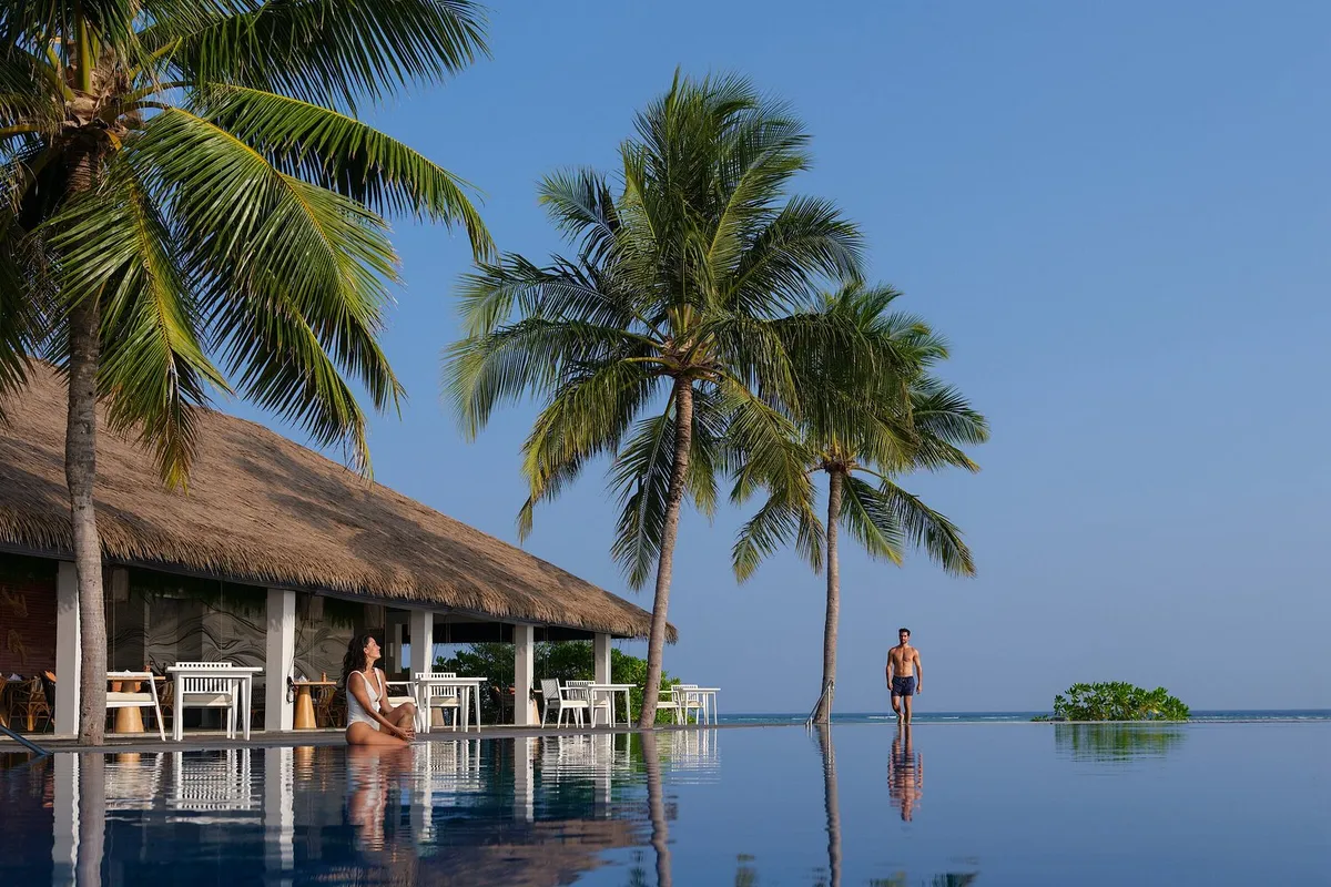 Noku Maldives, infinity pool with palm trees and thatched-roof pavilion at Kudafunafaru with mangrove islet on the horizon, Noonu Atoll, Maldives