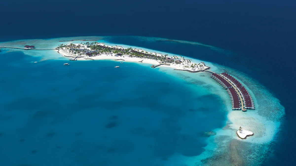 OBLU SELECT Lobigili, aerial, North Malé Atoll, Maldives, Daytime full-island aerial: the small crescent island geometry on the eastern North Malé Atoll edge with the red-roof overwater villa row curving along the southeast lagoon edge, sandbar at the southern tip, central island palm-canopied building cluster, reef bowl visible beneath turquoise lagoon.