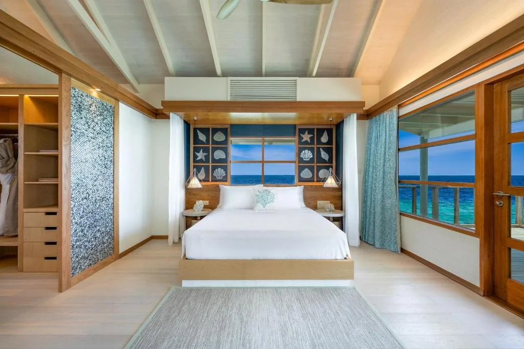 OBLU SELECT Lobigili, villa overwater, North Malé Atoll, Maldives, Water Villa bedroom interior: king bed with shell-display panel headboard (the Atmosphere brand signature framing starfish and conch in dark blue cabinetry panels), wooden ceiling slats, picture window to ocean, glass deck-door opening to the overwater step-down landing.