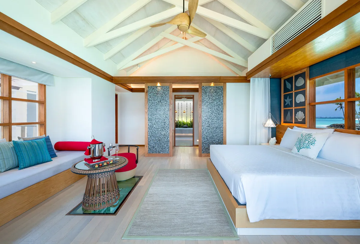 OBLU SELECT Lobigili, villa overwater, North Malé Atoll, Maldives, Water Villa with Pool wide interior: vaulted white-rafter ceiling with central palm-leaf fan, daybed banquette with blue and red cushions on the left wall, king bed with the same shell-panel headboard on the right, wooden floors, picture window framing the lagoon and beach beyond.