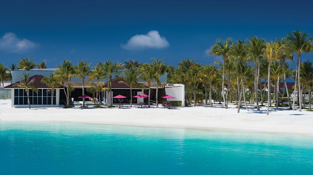 OBLU SELECT Lobigili, dining, North Malé Atoll, Maldives, Main dining pavilion viewed from the lagoon: glass-fronted modern pavilion with the property's signature magenta canvas umbrellas, palm-shaded along the white-sand beach edge, turquoise shallow lagoon foreground.