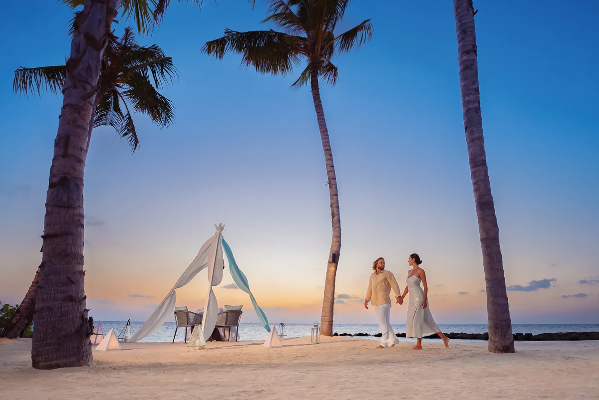 OBLU SELECT Lobigili, dining, North Malé Atoll, Maldives, Sunset private beach dinner staging: draped tipi-style canvas canopy with twin rattan chairs and lantern cluster on white sand, framed by leaning palms, dusk sky transitioning from orange horizon to deep blue overhead.