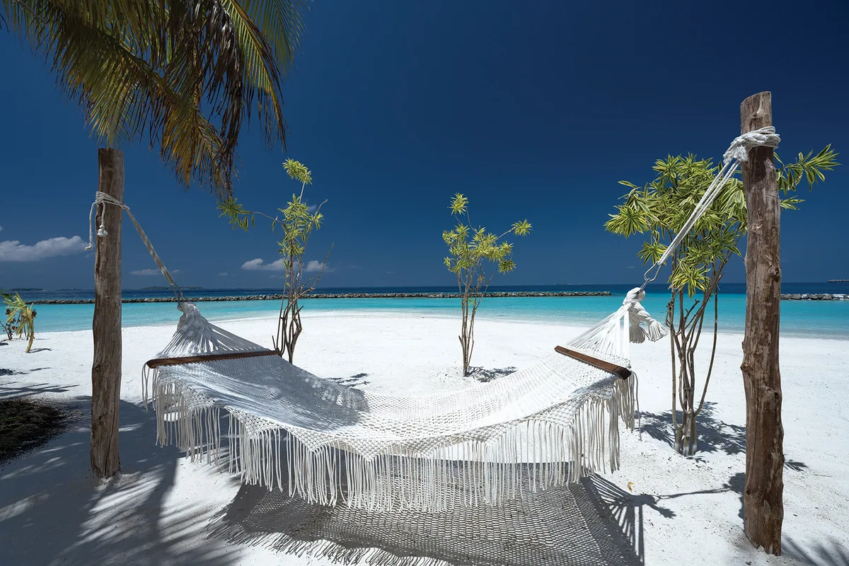 OBLU SELECT Lobigili, spa, North Malé Atoll, Maldives, Macramé hammock pavilion on the beach: hand-knotted off-white macramé hammock slung between two driftwood posts with rope binding, planted ornamental shrubs around, breakwater rocks and open lagoon backdrop, palm canopy upper frame.