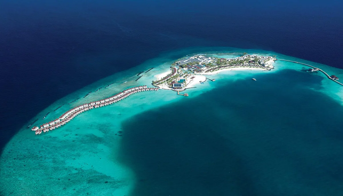 OBLU XPERIENCE Ailafushi, aerial, North Malé Atoll, Maldives, Full-island aerial: the elongated Ailafushi island geometry with the distinctive S-curve overwater villa row extending along the western reef edge (the property's architectural signature), central island with two large building clusters (main pavilion plus spa block), main pool flush with the southern beach edge, secondary jetty cluster at the eastern arm.