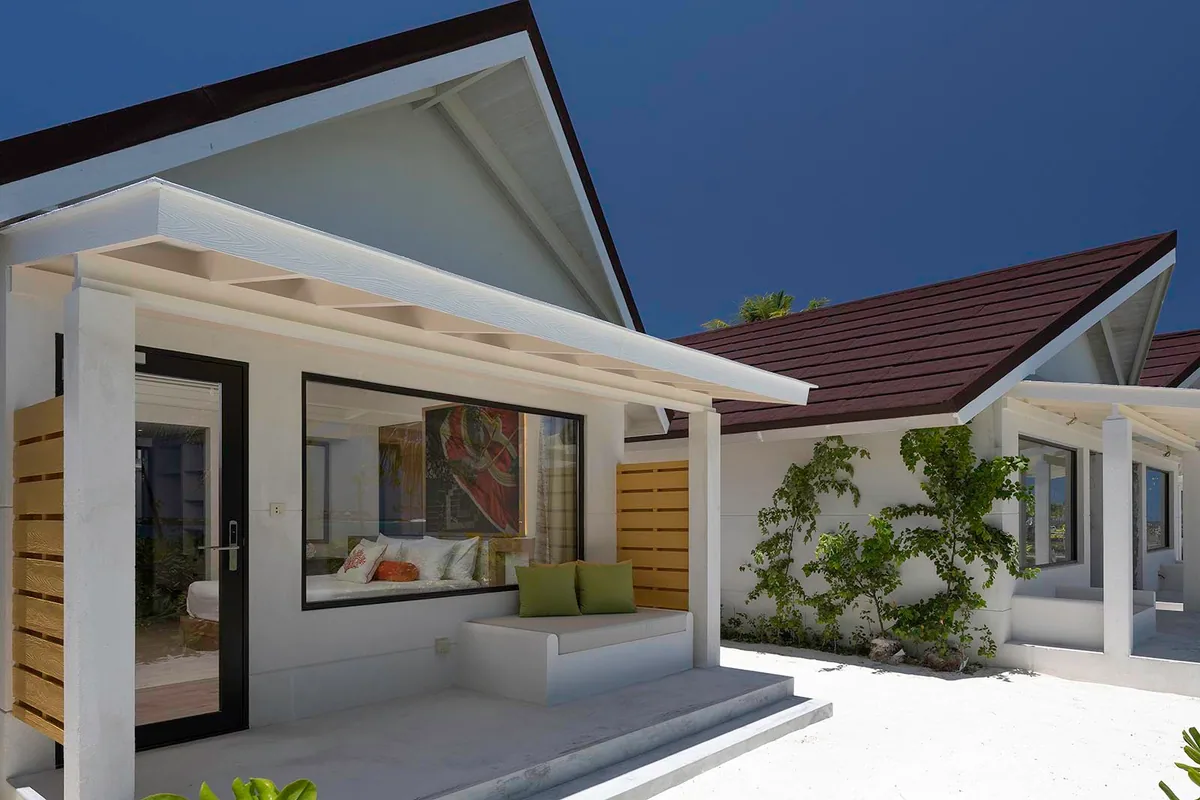 OBLU XPERIENCE Ailafushi, villa beach, North Malé Atoll, Maldives, Beach Villa exterior at dusk: contemporary white-walled pavilion with pitched red-tile roof (the Ailafushi cohort signature), entry porch with built-in bench under overhang, large picture window framing the king bed inside, yellow-louvered side privacy panels, planted shrub border between adjacent villas.