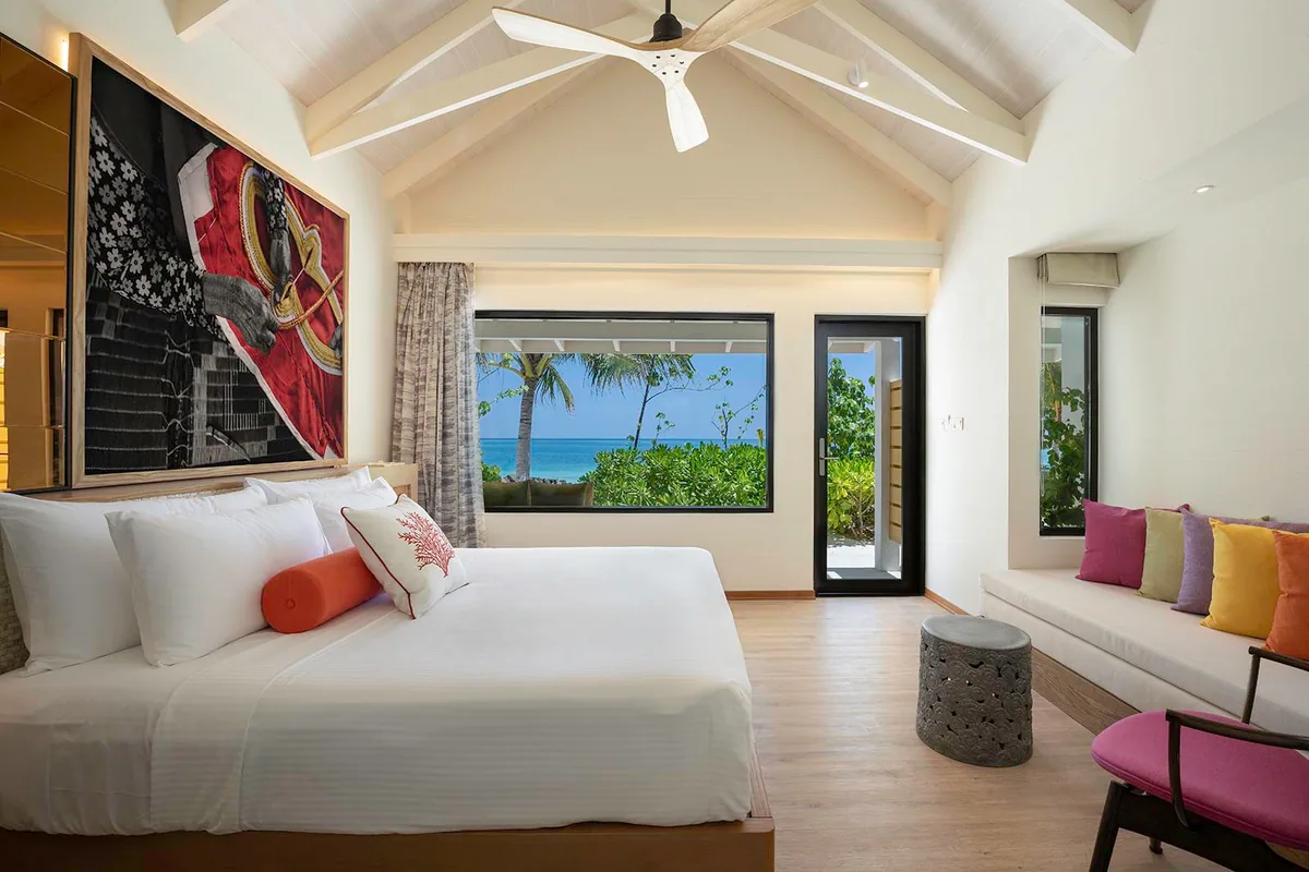 OBLU XPERIENCE Ailafushi, villa beach, North Malé Atoll, Maldives, Beach Villa bedroom interior: vaulted white-rafter ceiling with large rattan-blade fan, king bed dressed in white linens with coral-pattern pillow and orange bolster, oversized dark-framed monochrome art print of hands holding a coiled red fishing rope (Maldivian-photography signature), picture window framing palm and ocean, side daybed banquette with rainbow-pastel cushions, grey ornate stone side table.