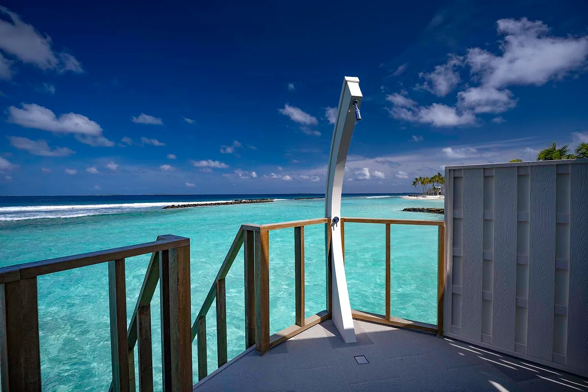 OBLU XPERIENCE Ailafushi, villa overwater, North Malé Atoll, Maldives, Water Villa deck configuration: timber-railed deck overhanging the lagoon with the property's curved white outdoor shower as the central feature, reef-line and breakwater visible across the turquoise water, island palms at the right edge.