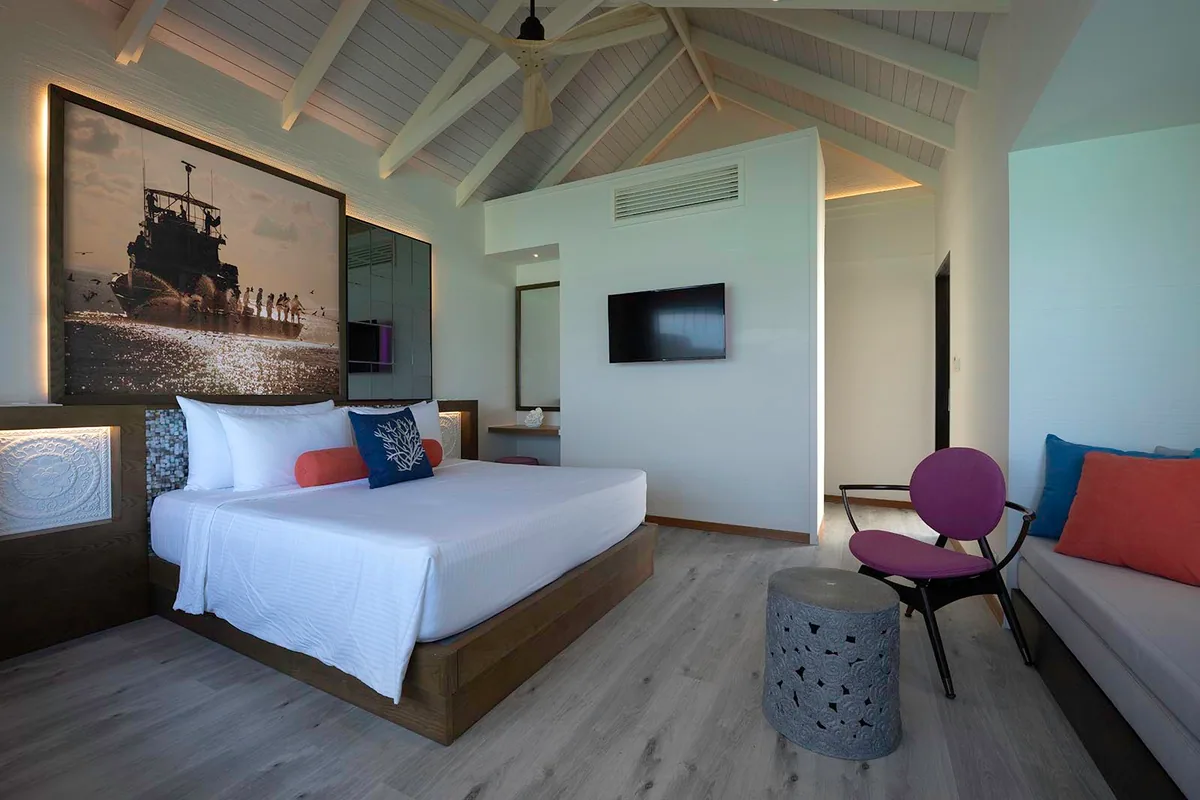 OBLU XPERIENCE Ailafushi, villa overwater, North Malé Atoll, Maldives, Water Villa bedroom interior: vaulted white-rafter ceiling with central ceiling fan, king bed on a dark-wood platform with ornate carved side panels and recessed accent lighting, oversized photographic art print of dhoni fishermen pulling a boat ashore (Maldivian-photography signature continuing the brand thread across villa categories), wall-mounted television, side daybed with orange-and-blue cushions.