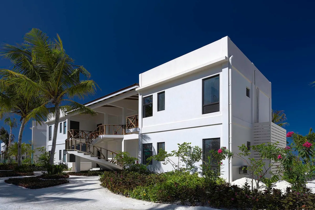 OBLU XPERIENCE Ailafushi, villa beach, North Malé Atoll, Maldives, Ocean View Family Room building exterior: two-storey modern white-walled flat-roof block (architecturally distinct from the single-storey pitched-roof beach villas), exterior staircase to the upper-floor balcony, palm trees flanking, planted ground border, distinct family-cohort building typology supporting the multi-bedroom inventory.