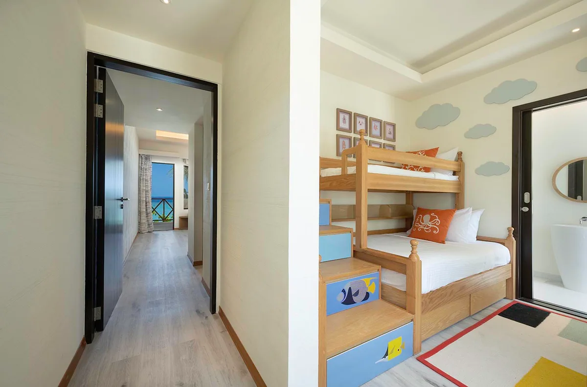 OBLU XPERIENCE Ailafushi, villa beach, North Malé Atoll, Maldives, Ocean View Family Room dedicated children's bunk area: built-in wooden bunk bed with octopus and sea-creature appliqué pillows on white linens, painted blue cloud wall decals above the bunks, ship-ladder steps with tropical-fish painted risers, framed nautical-themed prints, glimpsed main bedroom with ocean view through the open corridor door, the family-all-inclusive structural distinctive within the room category.