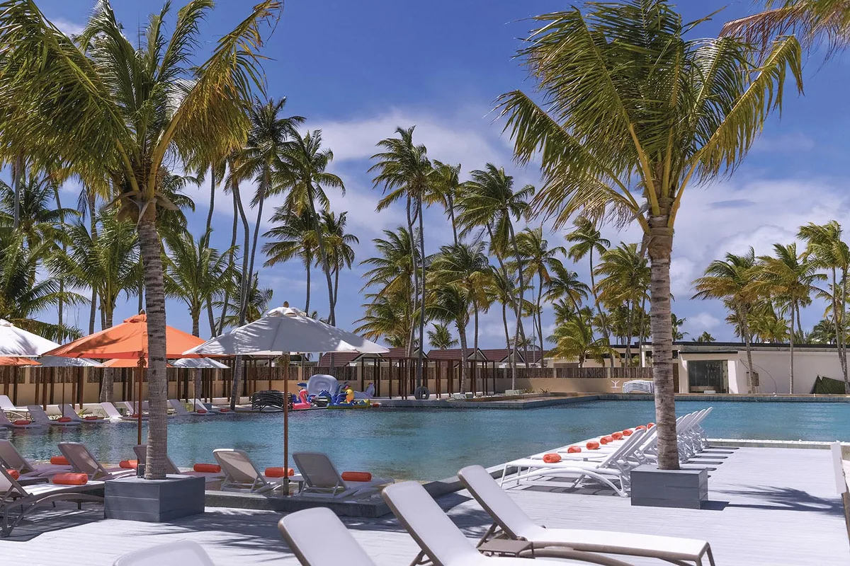 OBLU XPERIENCE Ailafushi, setting, North Malé Atoll, Maldives, Main central pool: large rectangular pool with white loungers and orange canvas umbrellas around the perimeter, family pool toys floating in the shallow end, dense palm canopy filtering the overhead light, the value-all-inclusive Sunset Pool Bar pavilion glimpsed beyond.