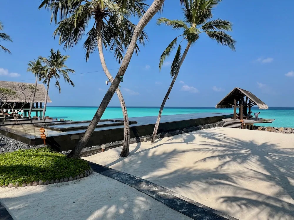 Dark-tile main pool with palm fringe, thatched gazebo and lagoon backdrop at One&Only Reethi Rah.