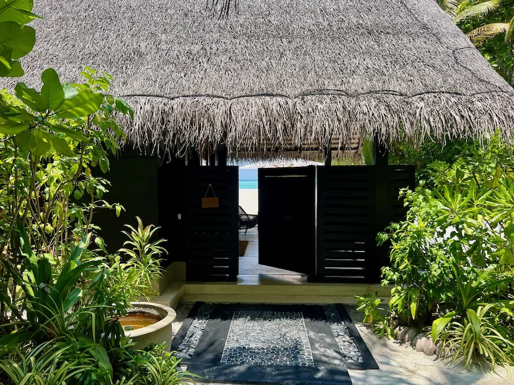 One&Only Reethi Rah Beach Villa entrance with dark wood doors, thatched roof and a hallway through to the beach.