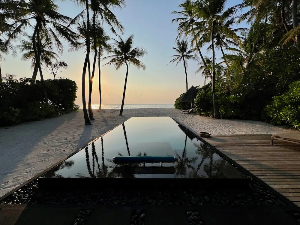 Beach Villa dark-tile reflection pool at sunset with palm silhouettes and a thatched gazebo at One&Only Reethi Rah.