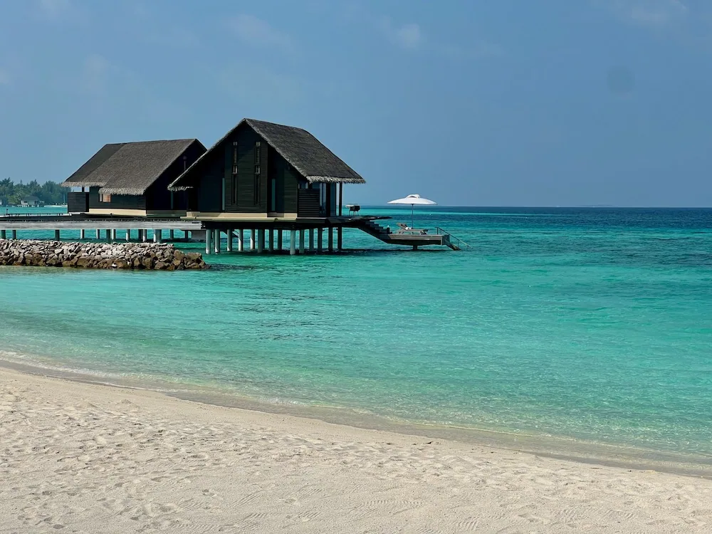 Over-water pavilions with dark gabled thatched roofs on stilts above turquoise lagoon, rocky breakwater foreground at One&Only Reethi Rah.