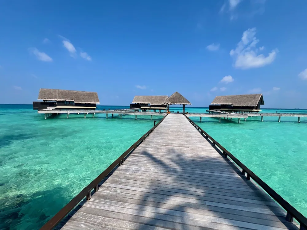 Boardwalk leading axially down to three thatched-roof Water Villas over emerald lagoon at One&Only Reethi Rah.