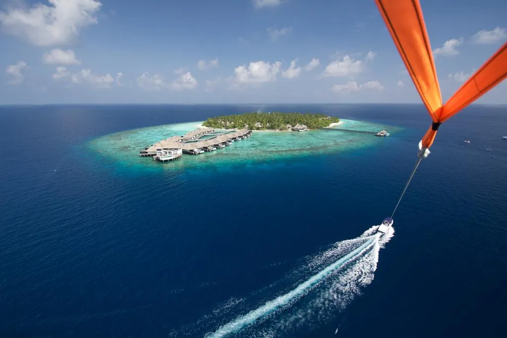 Outrigger Konotta Maldives Resort, aerial, Gaafu Dhaalu Atoll, Maldives, Outrigger Konotta Maldives Resort, the Konotta island geometry in Gaafu Dhaalu Atoll with 53-villa Hawaiian-DNA boutique luxury configuration: parasail-mounted aerial showing the palm-covered island ringed by the over-water villa cluster on the southern lagoon edge, with a speedboat trail crossing the deep-water channel; orange parasail wing visible upper-right (paragliding viewpoint).