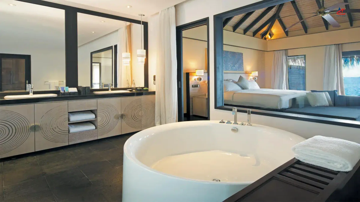 Outrigger Konotta Maldives Resort, villa overwater, Gaafu Dhaalu Atoll, Maldives, Ocean Pool Suite over-water configuration bathroom interior: double-vanity counter with paired wood-grain cabinets, freestanding circular soaking tub, full-length framed mirror separating the bath area from the bedroom which is visible through the doorway with its made bed and corner window onto the lagoon.