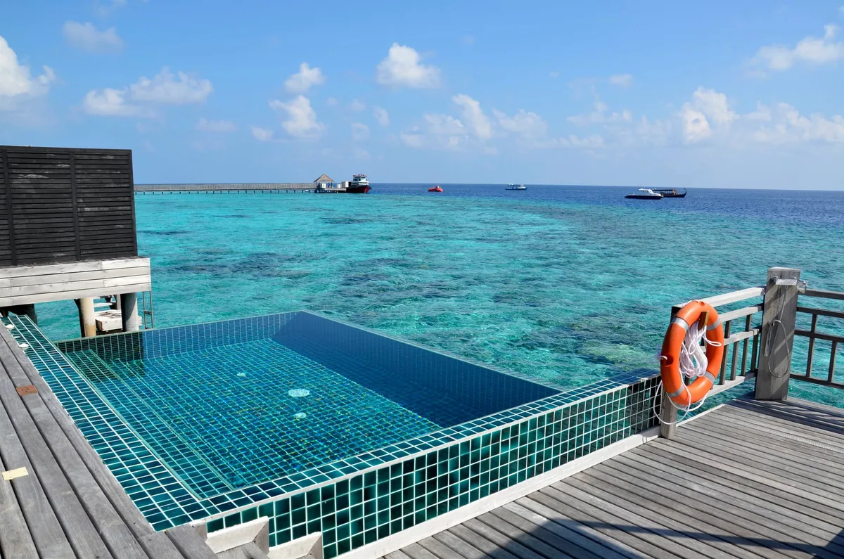 Outrigger Konotta Maldives Resort, villa overwater, Gaafu Dhaalu Atoll, Maldives, Ocean Pool Villa private deck: infinity-edge plunge pool with mosaic-blue tile bottom stepping into the lagoon, weathered-teak decking, life-ring on the railing (operator safety signage), house reef visible through the clear water, speedboats moored on the deep-channel side.