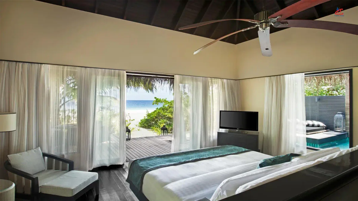 Outrigger Konotta Maldives Resort, villa beach, Gaafu Dhaalu Atoll, Maldives, Beach Pool Suite at the perimeter beach pool configuration with Hawaiian-DNA aloha-spirit aesthetic elements: contemporary bedroom interior with floor-to-ceiling glazing onto the beach-back vegetation and lagoon view, ceiling-fan, dark hardwood floor, plunge-pool deck visible through the side glazing.
