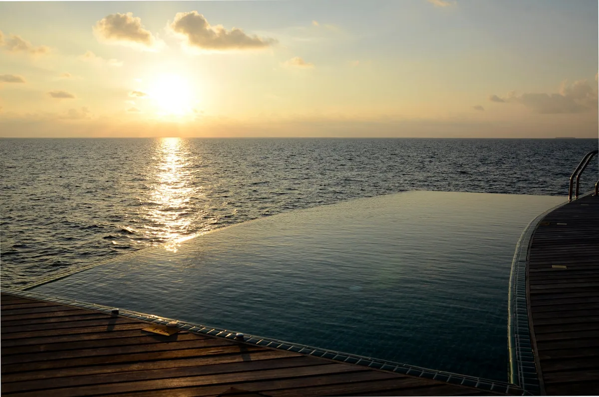 Outrigger Konotta Maldives Resort, villa overwater, Gaafu Dhaalu Atoll, Maldives, Ocean Pool Villa private infinity pool at sunset, the western open-ocean horizon catching the gold light, dark teak deck with pool-side ladder, no other property in view (the southernmost outer-atoll position visible).