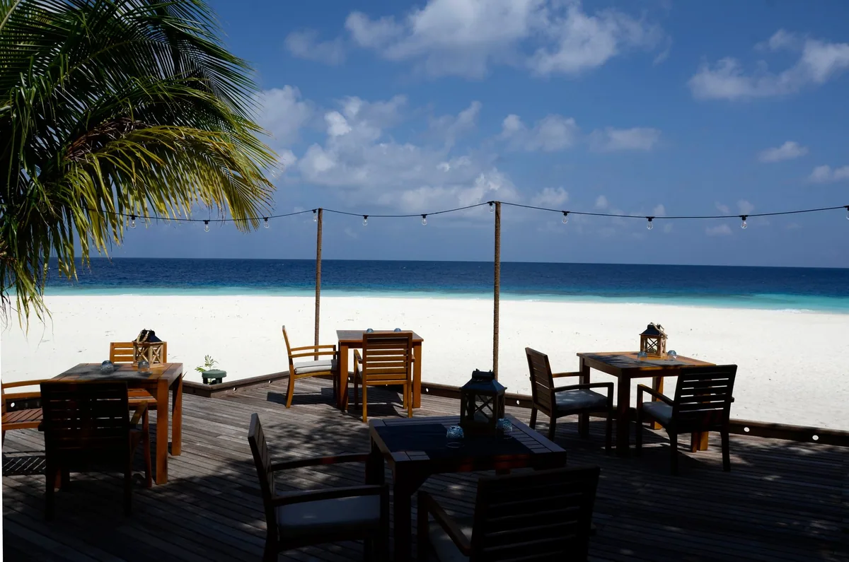Outrigger Konotta Maldives Resort, dining, Gaafu Dhaalu Atoll, Maldives, Beachfront dining deck with string-light pergola posts running parallel to the shore, wood-and-cane chairs at four-top tables set with brass storm lanterns, palm canopy shading the foreground, white sand and the turquoise lagoon framed beyond the deck rail.