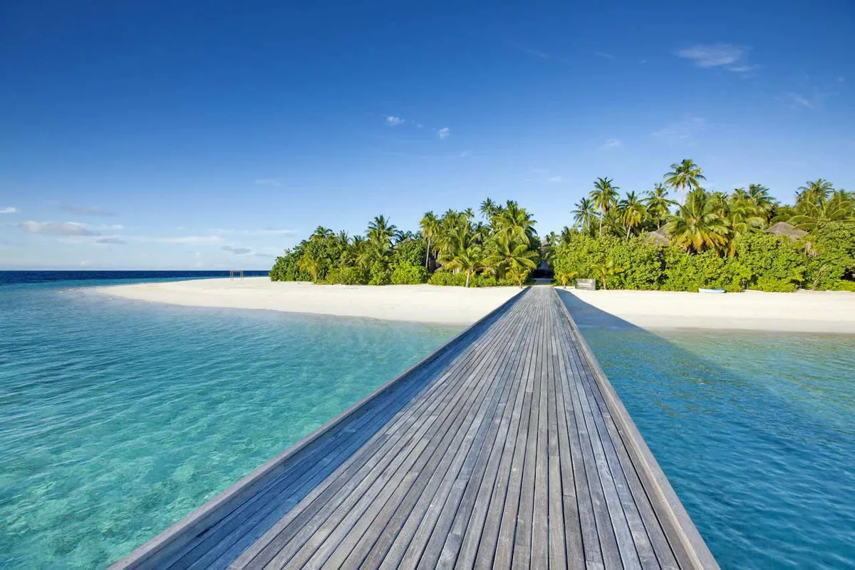 Outrigger Konotta Maldives Resort, setting, Gaafu Dhaalu Atoll, Maldives, The Konotta jetty bridge from POV at the lagoon-side mouth: a single teak-plank boardwalk running straight onto the palm-thick island, white sandbar tip flanking the lagoon-shallow side, thatched villa roofs visible through the palm canopy at the landfall.