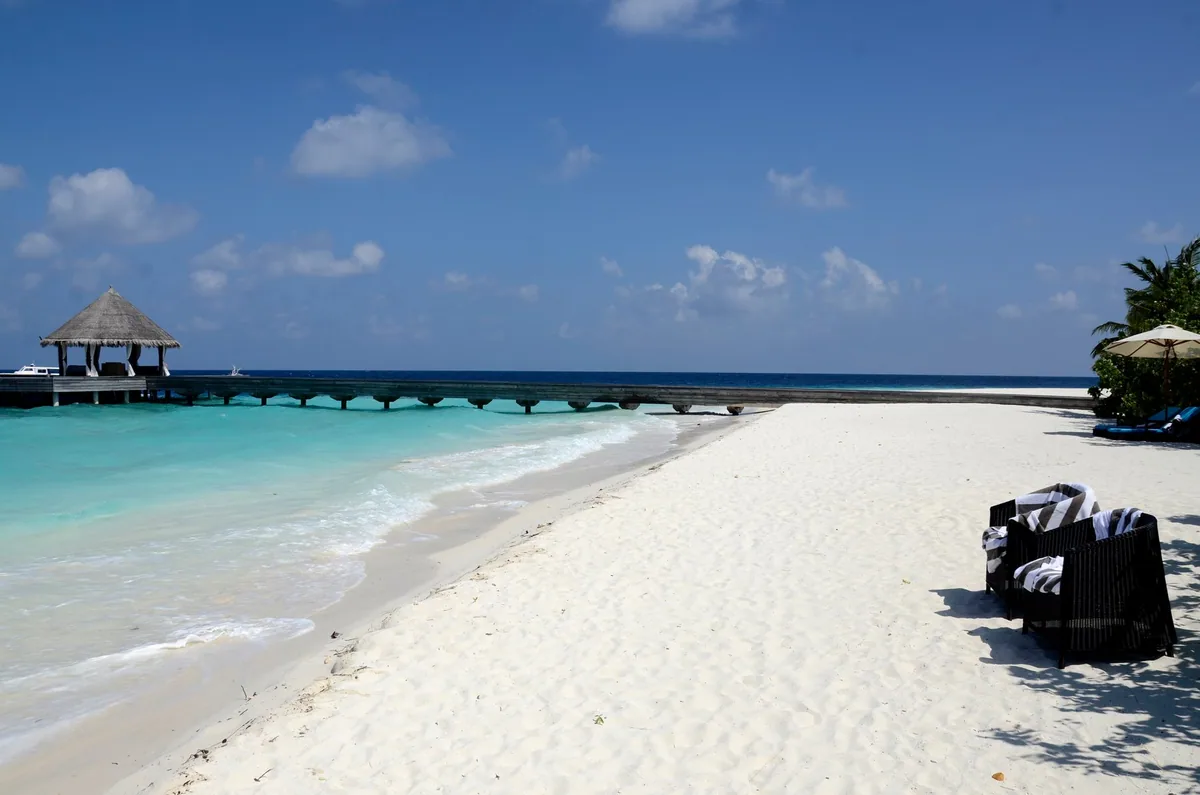 Outrigger Konotta Maldives Resort, setting, Gaafu Dhaalu Atoll, Maldives, Konotta arrival side from the beach: the parallel jetty bridge ending at a thatched-pyramid welcome pavilion at the far end, rattan beach chairs draped with Outrigger striped towels in the foreground, lone parasol shading them.