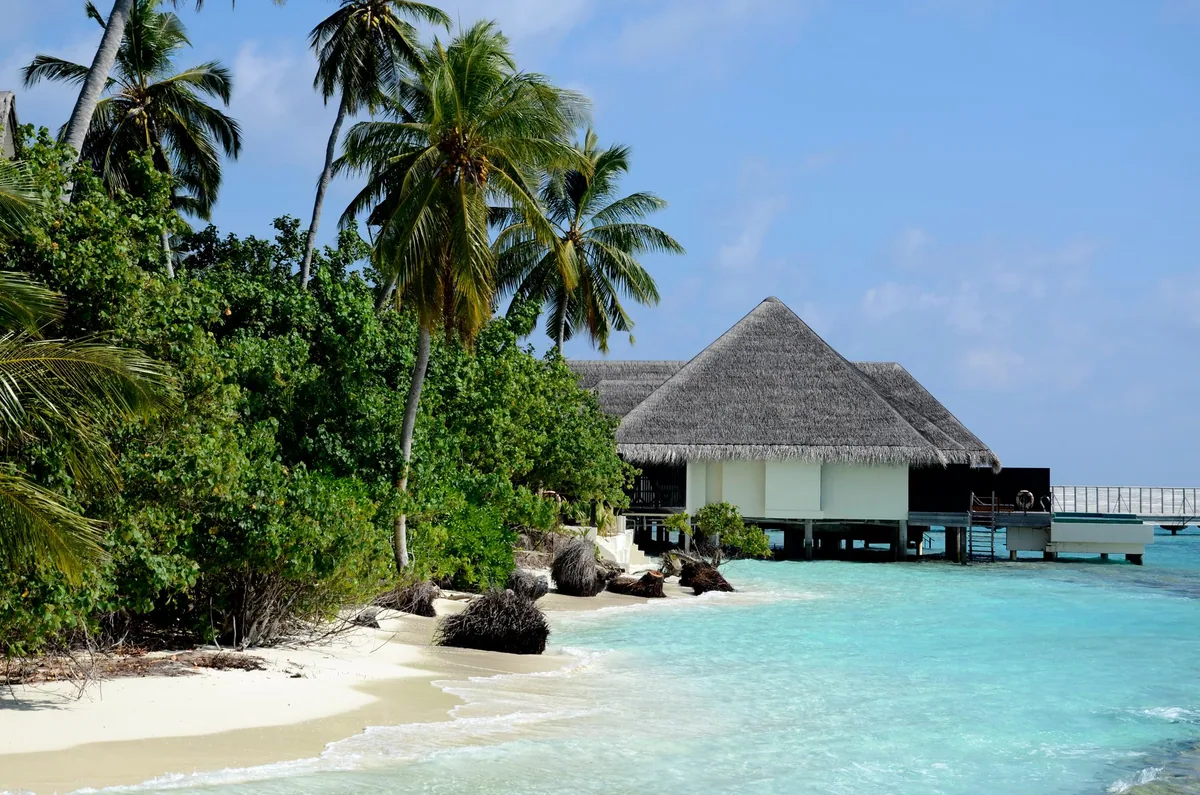 Outrigger Konotta Maldives Resort, spa, Gaafu Dhaalu Atoll, Maldives, Navasana Spa lagoon-edge pavilion: thatched pyramidal roof with dark timber facing, palm canopy and rounded shoreline boulders flanking the entrance, the overwater villa walkway visible behind the pavilion, calm turquoise lagoon in the foreground.