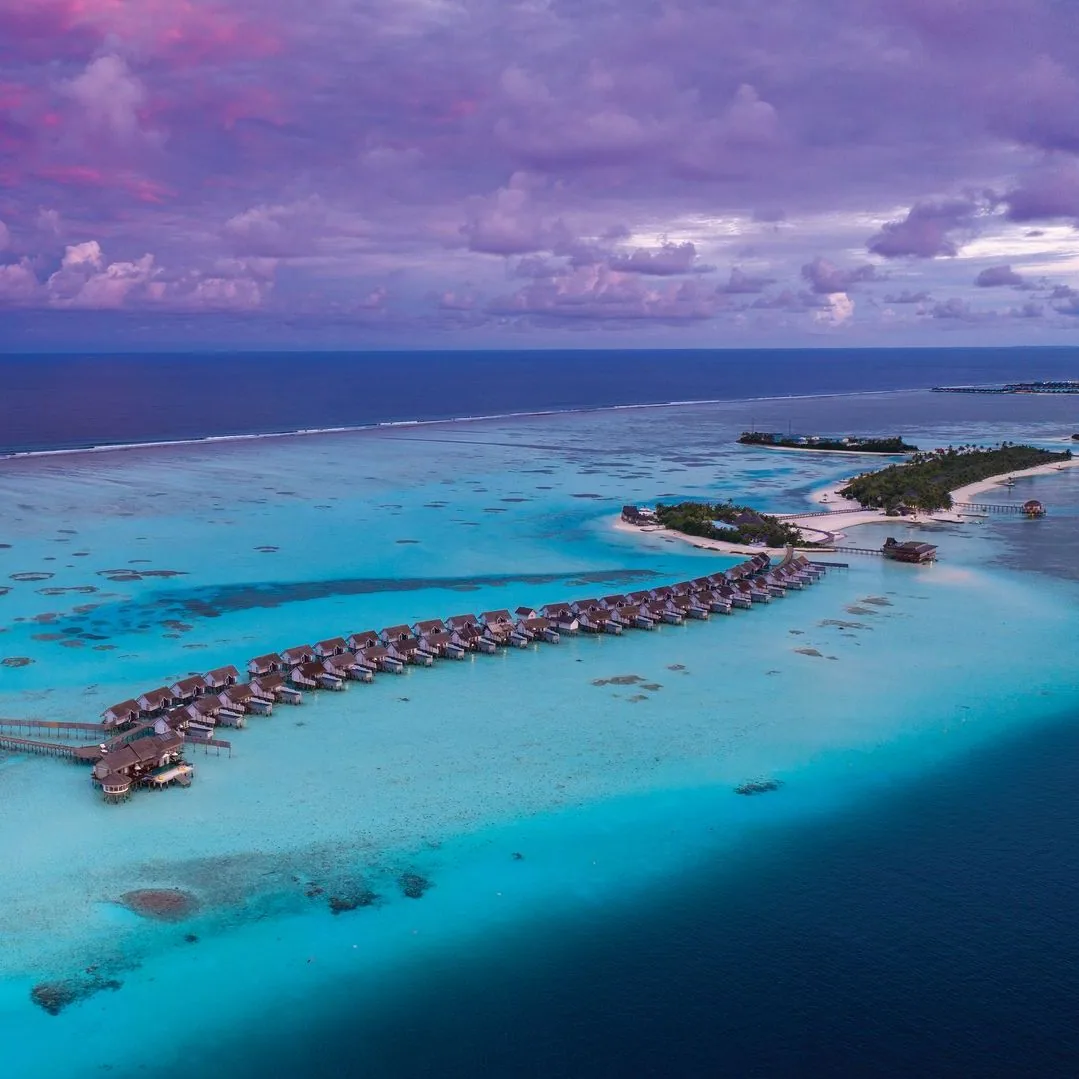 OZEN LIFE MAADHOO, aerial, South Malé Atoll, Maldives, OZEN LIFE MAADHOO sunset drone aerial: the curving over-water villa row extending into the South Malé Atoll lagoon, violet-pink twilight sky overhead, the main Maadhoo island visible at the right edge with its secondary jetty pavilion, deep-water channel and reef edge in the foreground.
