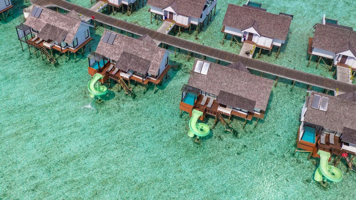 OZEN LIFE MAADHOO, aerial, South Malé Atoll, Maldives, Aerial of the Wind Villa row with three green spiral waterslides visible spiralling from villa decks down into the lagoon, peaked shingle roofs along the jetty, the boardwalk path between, the documentary face of the OZEN green-slide tier across the inventory.