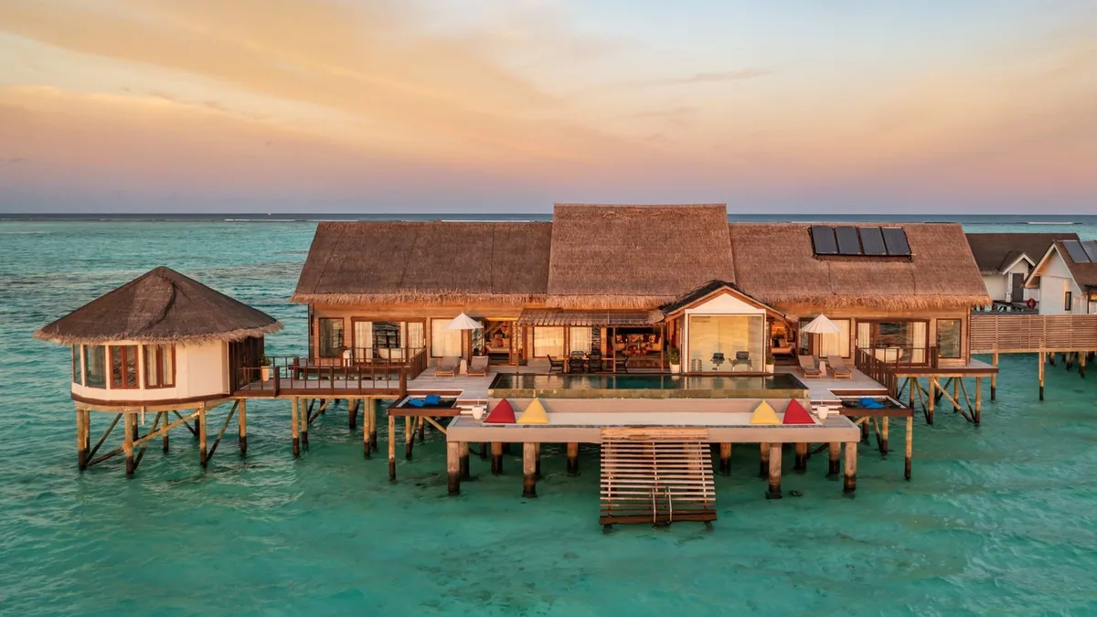 OZEN LIFE MAADHOO, aerial, South Malé Atoll, Maldives, Multi-room overwater suite in eye-level sunset: thatched main pavilion with a round-thatched side cabana, a large infinity pool with four bean-bag loungers along the dock edge, a stepped jetty descending into the lagoon, the sky painted soft pink and gold.
