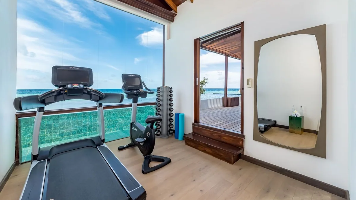 OZEN LIFE MAADHOO, wellness, South Malé Atoll, Maldives, Overwater fitness pavilion: treadmill and stationary bike positioned at a floor-to-ceiling glass wall over the lagoon, dumbbell rack and yoga rollers beside, the villa pool visible just below the deck rail, the ocean horizon beyond.