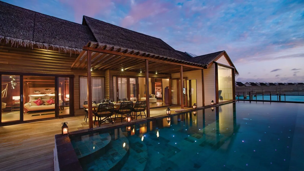 OZEN LIFE MAADHOO, dining, South Malé Atoll, Maldives, Multi-room overwater suite at twilight: pergola-covered private outdoor dining setup for six on the wooden deck, candle lanterns ringing the table, the infinity pool reflecting pink and blue light specks, the villa living room glowing through the glass wall behind, the sky brushed with violet stripes.